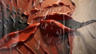 A single red poppy painted in thick palette-knife oils on canvas, the petals ridged so they catch warm studio light, with a small painting knife laying a final red stroke.